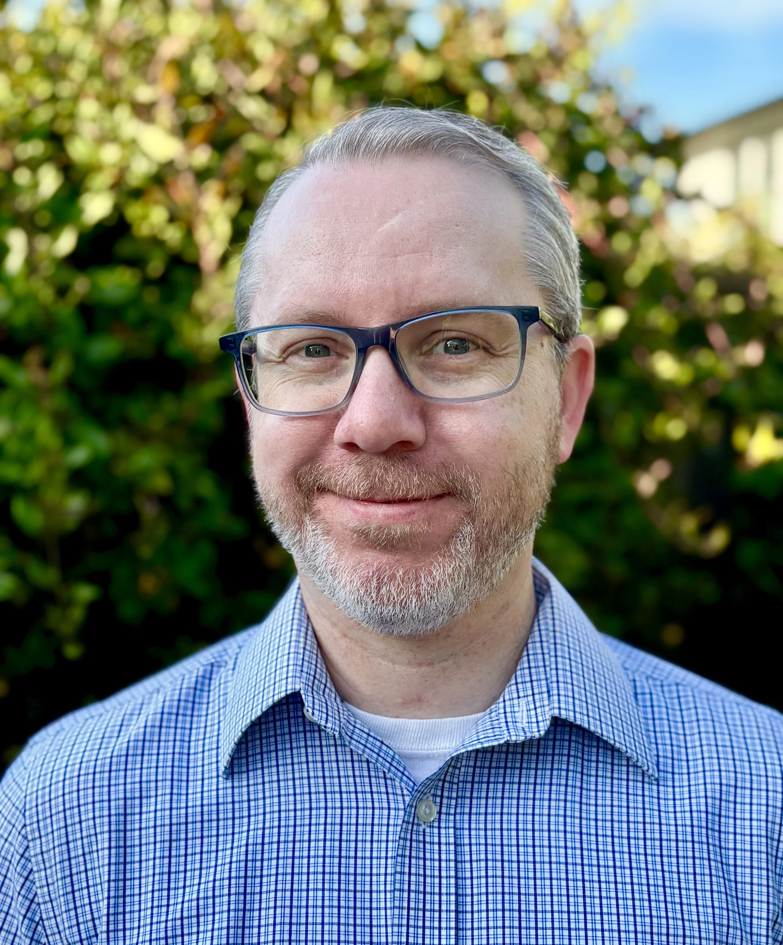 Close-up of a middle-aged man with gray hair and beard, wearing glasses and a blue checkered shirt, smiling outdoors with a blurred background of green foliage and blue sky.