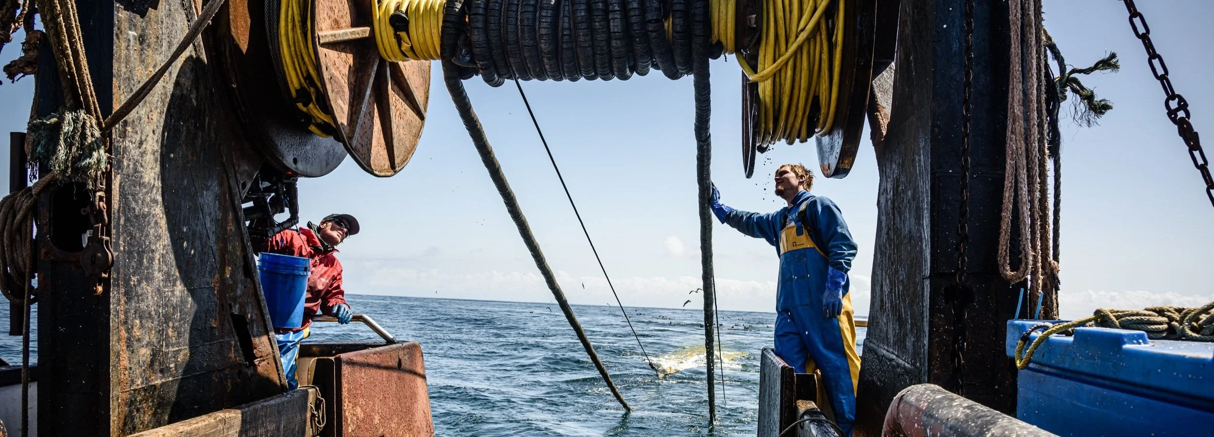 Two crew members on a research vessel deploying a large net into the ocean, with one person in a red jacket and the other in a blue and yellow suit, under a clear sky.