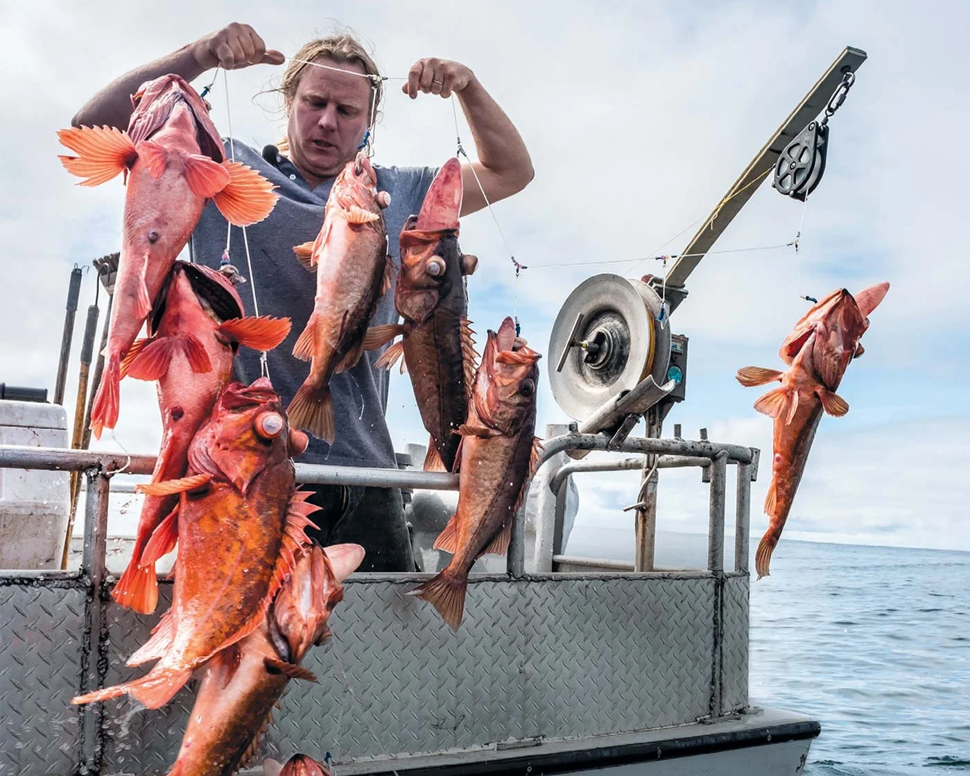 A man on a boat holding up a string of freshly caught red fish, with a meat slicer on the boat and open water in the background.