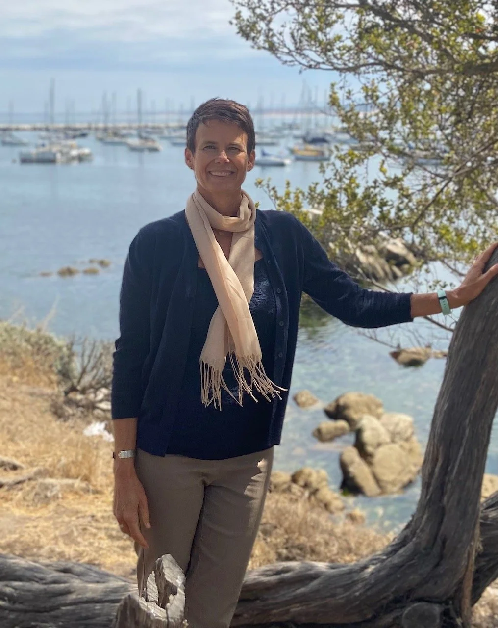 A woman standing outdoors near the water, with boats anchored in the harbor behind her and a tree in the foreground.