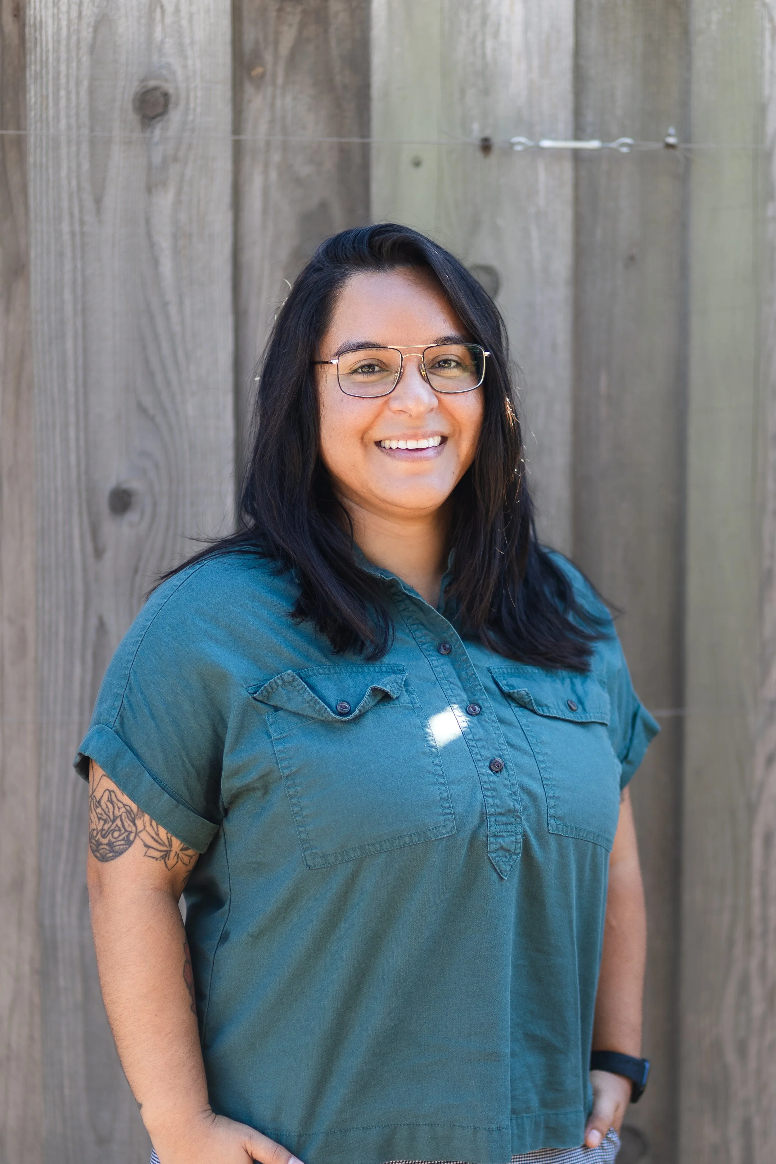 A woman with dark hair, glasses, and a tattoo on her arm, smiling and wearing a blue button-up shirt, standing in front of a wooden fence.