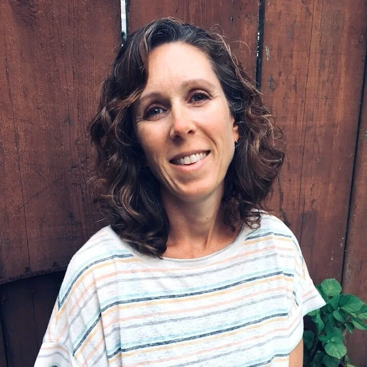 A woman with curly brown hair and a light striped t-shirt standing outdoors in front of a wooden fence and some green plants.