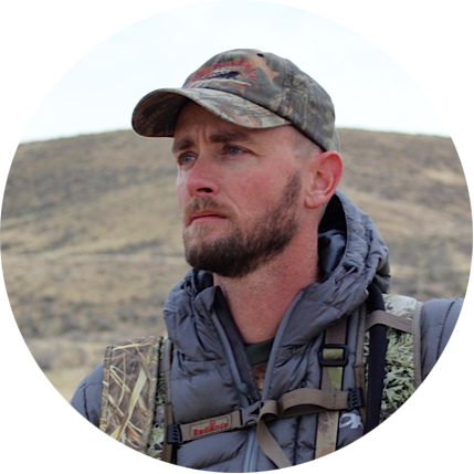 Man wearing camouflage baseball cap and outdoor gear outdoors in a rural landscape.