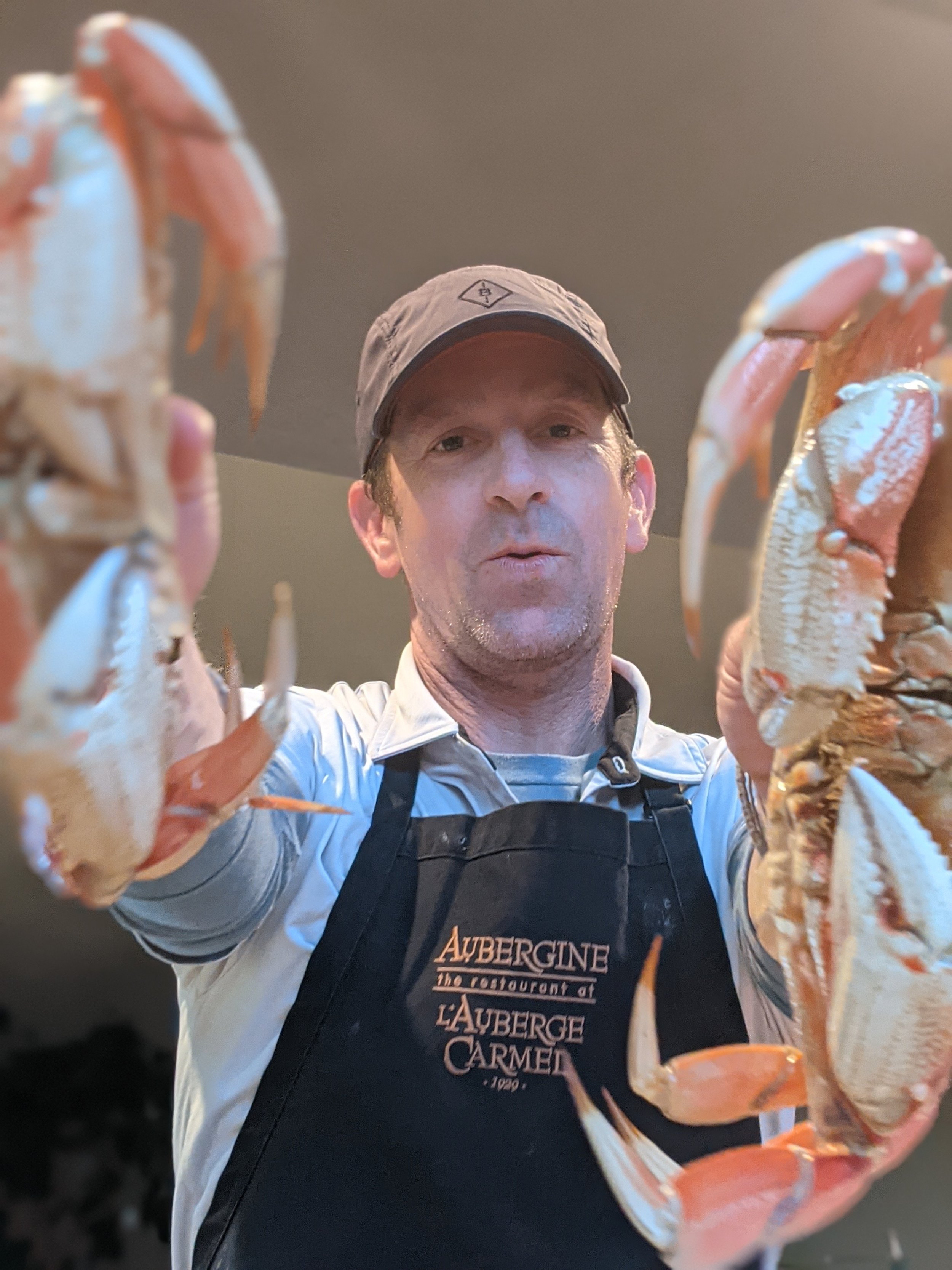 Man wearing a black apron with 'Auberge Carm' written on it, holding two cooked lobsters in front of the camera, with a neutral background.