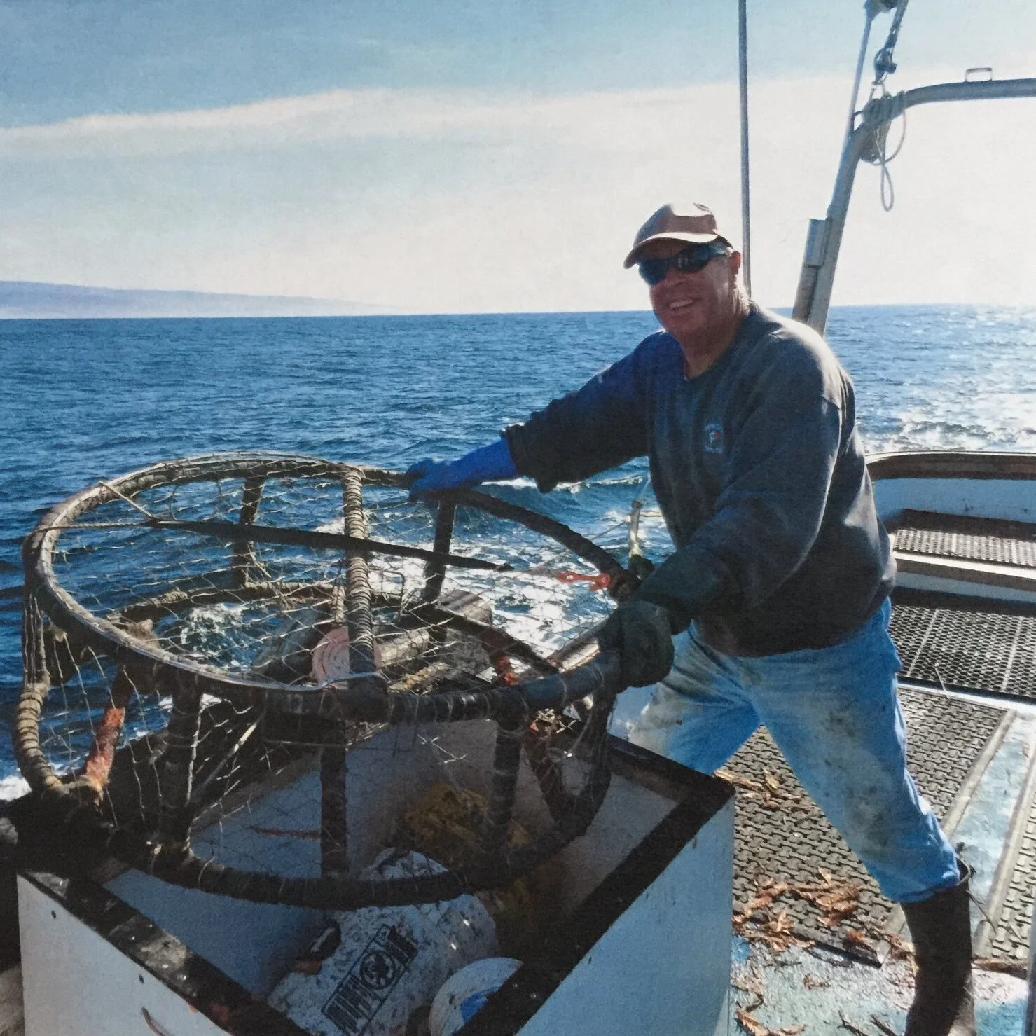 A man wearing a hat, sunglasses, and waterproof clothing is on a boat, smiling as he handles a large lobster trap. The boat is out on the ocean with a distant horizon visible in the background.