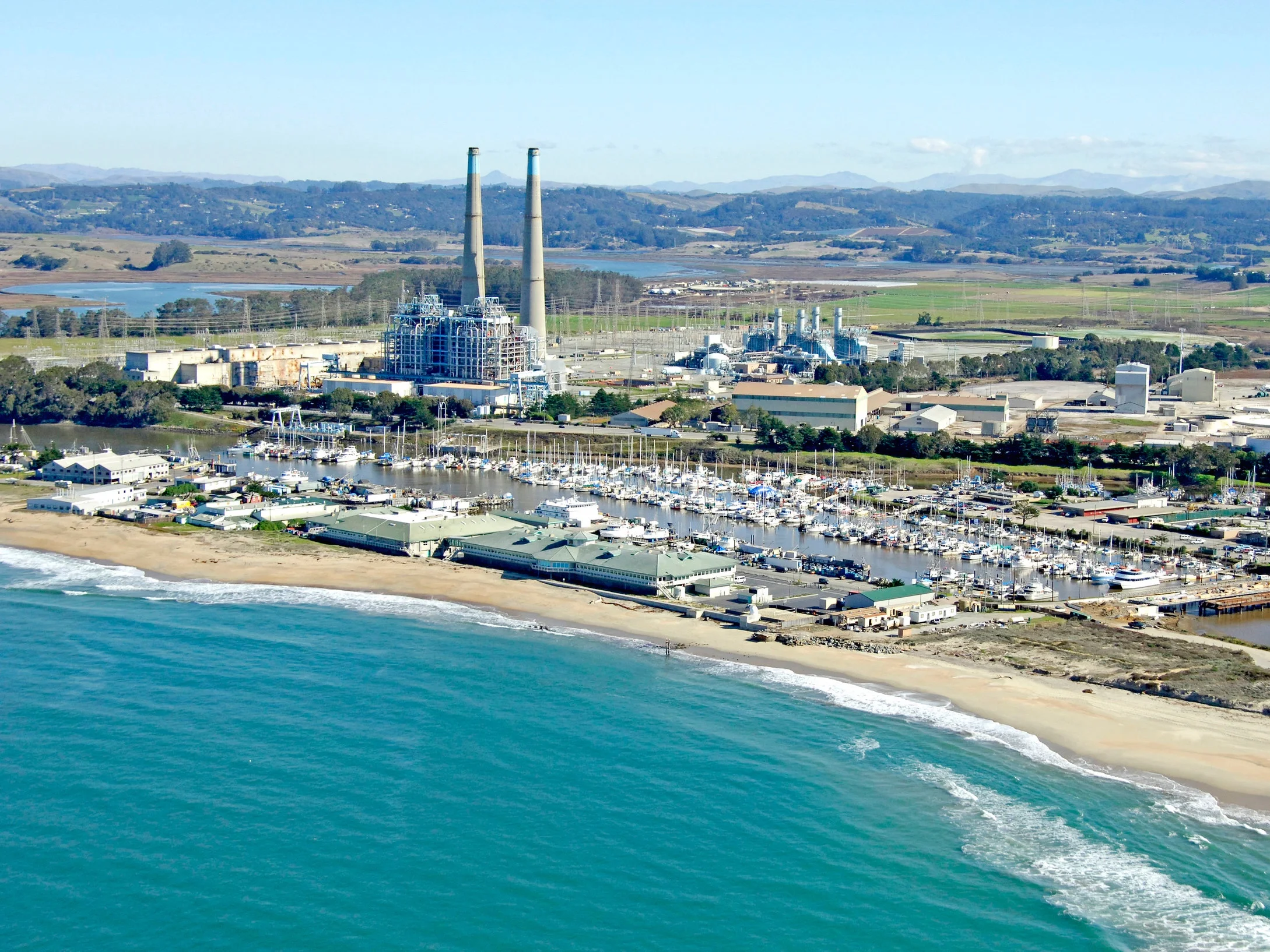 An aerial view of a coastal area featuring a beach, a marina with boats, industrial buildings, and smokestacks, with fields, lakes, and mountains in the background.