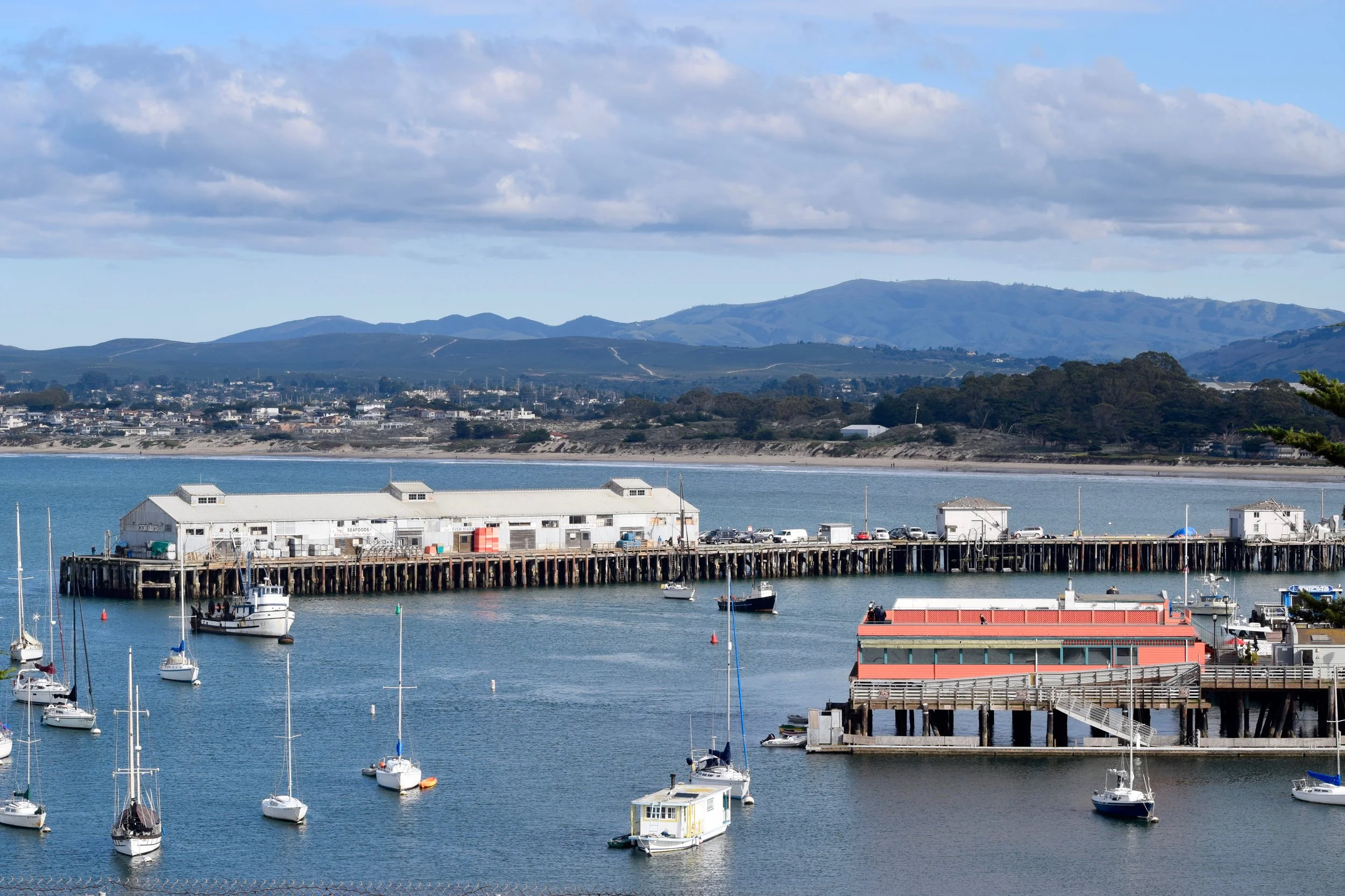 A coastal scene featuring boats docked in a marina, a pier with small buildings, and rolling hills in the background under a partly cloudy sky.