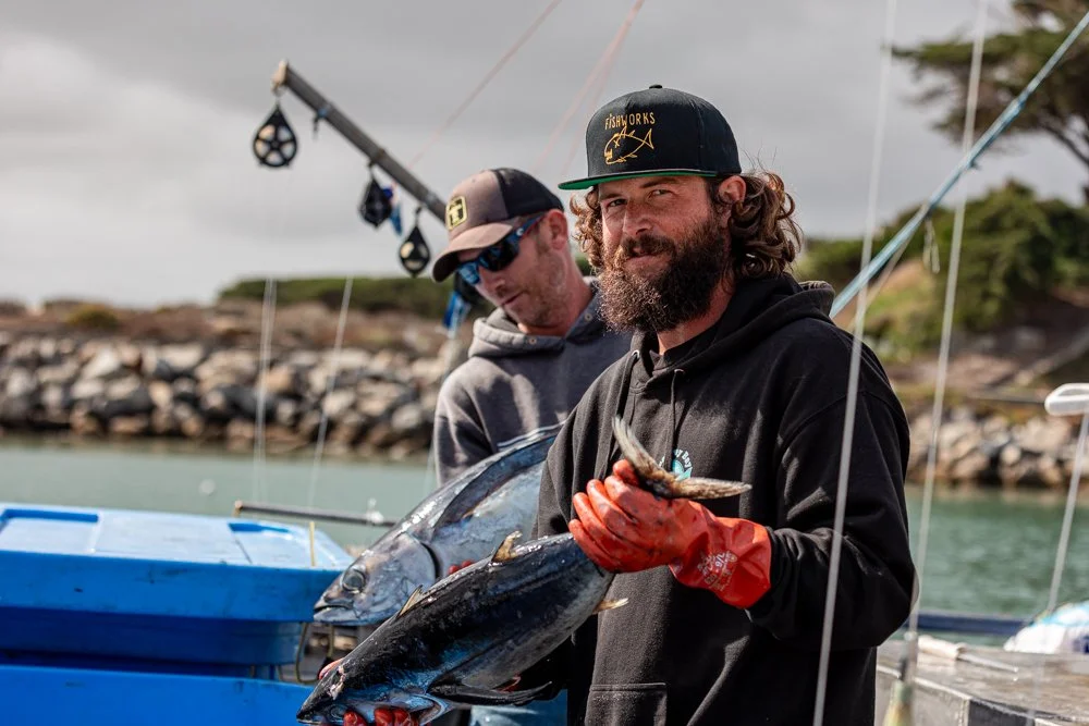A man with a beard and wearing a black cap, black hoodie, and red gloves poses with a fish he caught, while another person in a gray hoodie and cap stands beside him on a boat.
