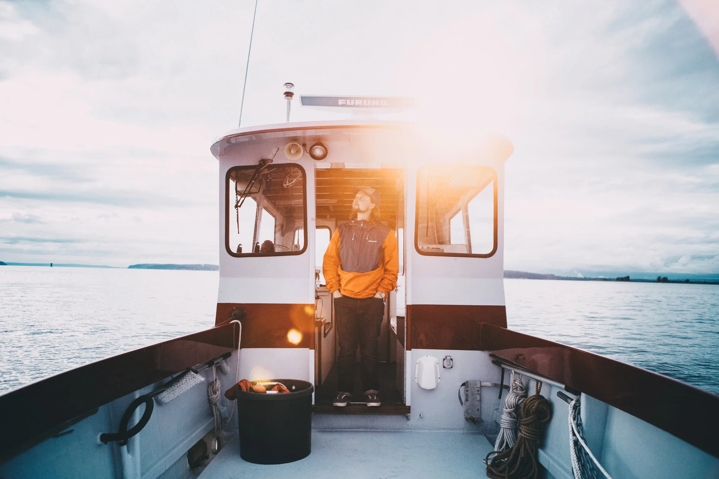 Man standing on the deck of a boat, wearing a yellow and gray jacket, looking out at the water with cloudy skies.