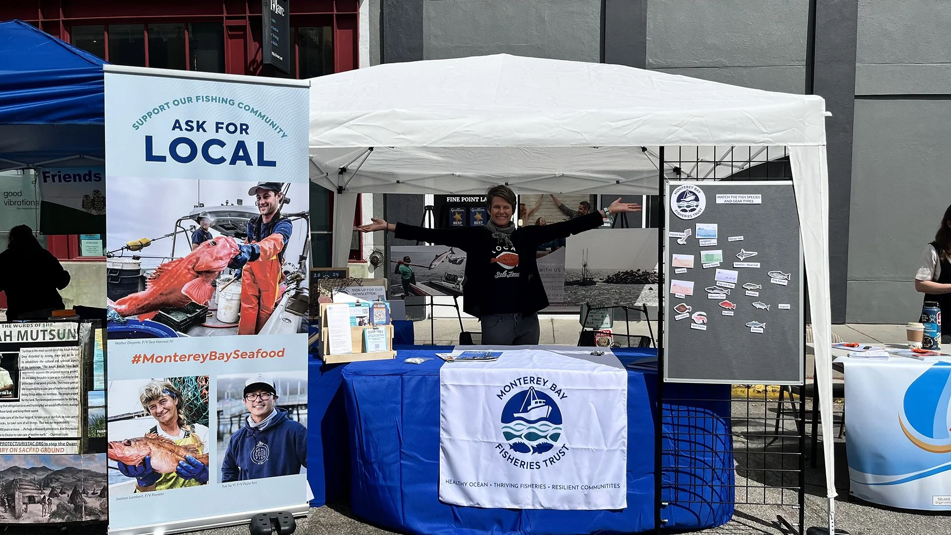 A woman standing under a white canopy tent at a Monterey Bay Fisheries Trust event, smiling with arms outstretched. The tent displays informational posters about fishing and local seafood, with a large sign encouraging support for local fishing community. There are chairs, tables, and a board with fish species and gear types on the side.