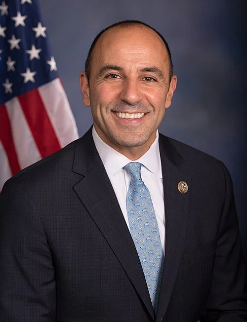 A smiling man in a suit with a blue tie, standing in front of an American flag and a dark background.