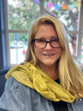Woman with blonde hair and glasses smiling indoors, wearing a denim jacket and a yellow shawl.