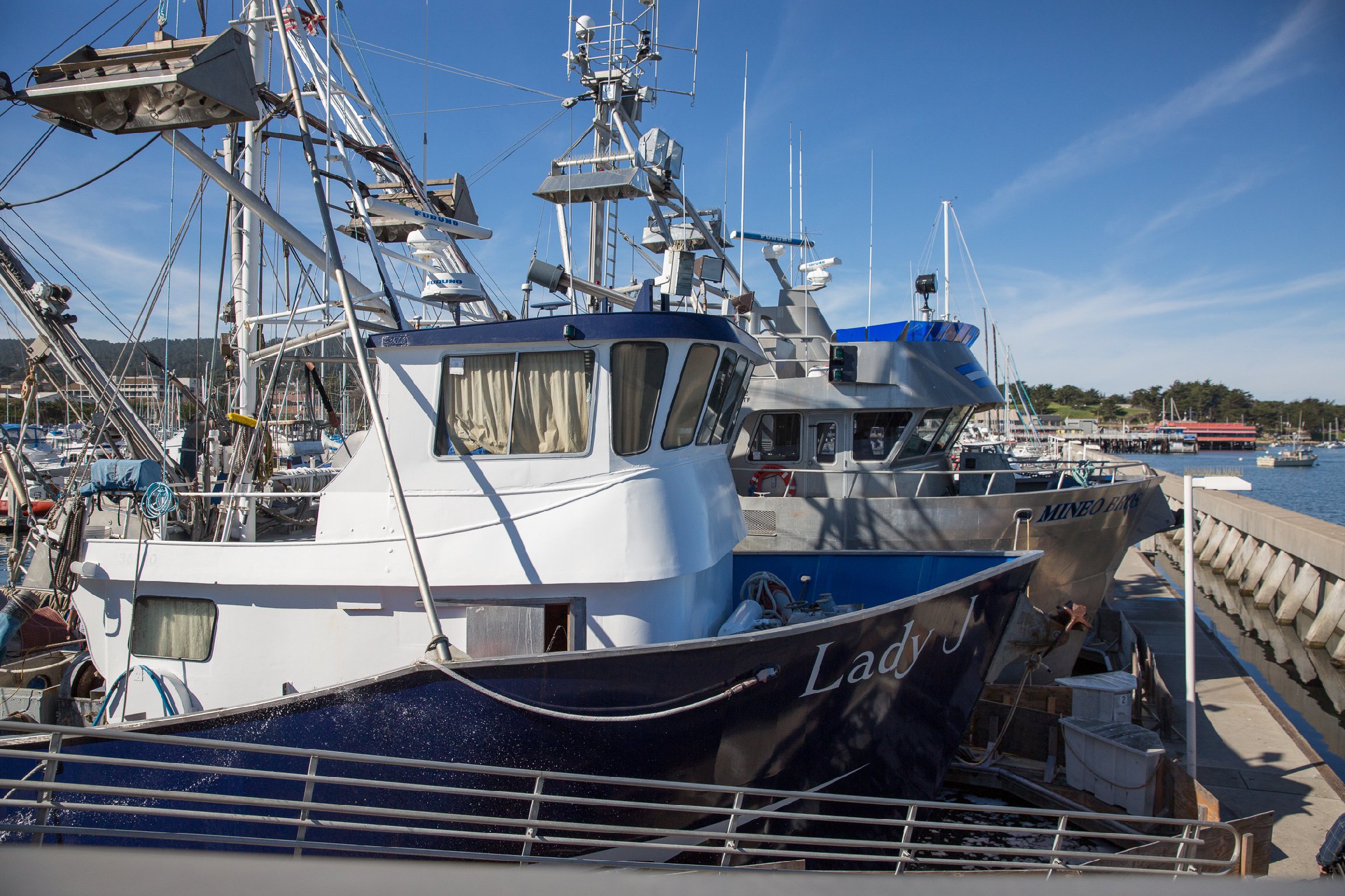 A docked yacht with various navigation and communication equipment on the upper deck, adjacent to a marina with other boats and a hilly landscape in the background, under a partly cloudy blue sky.