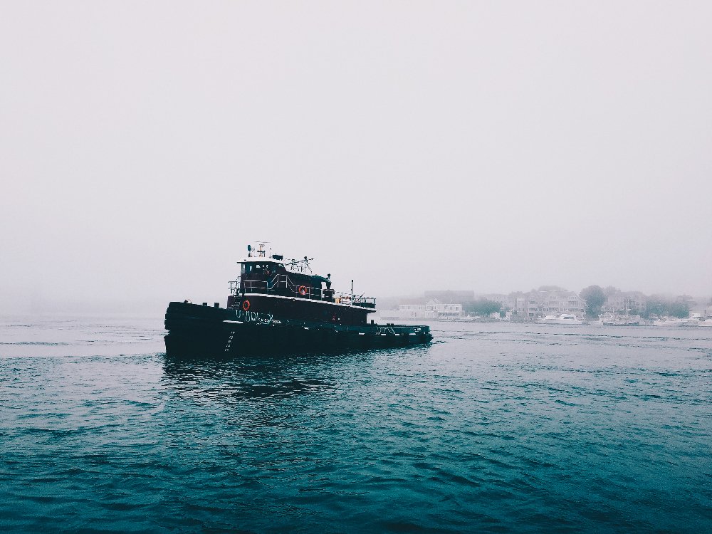 A black tugboat moving through calm water with foggy shoreline and buildings in the background.
