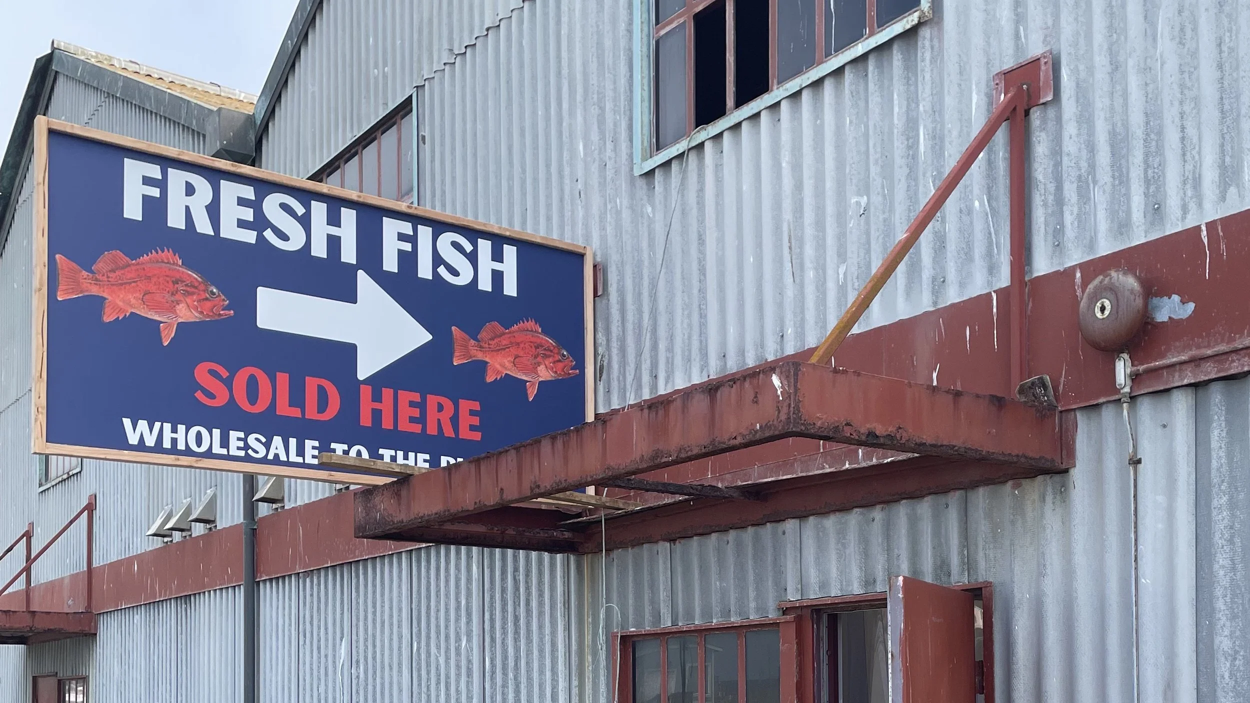 A sign for a fish market with two red fish images and the text 'FRESH FISH SOLD HERE WHOLESALE TO THE PUBLIC' on a blue background, mounted on a weathered building with corrugated metal siding and red trim.