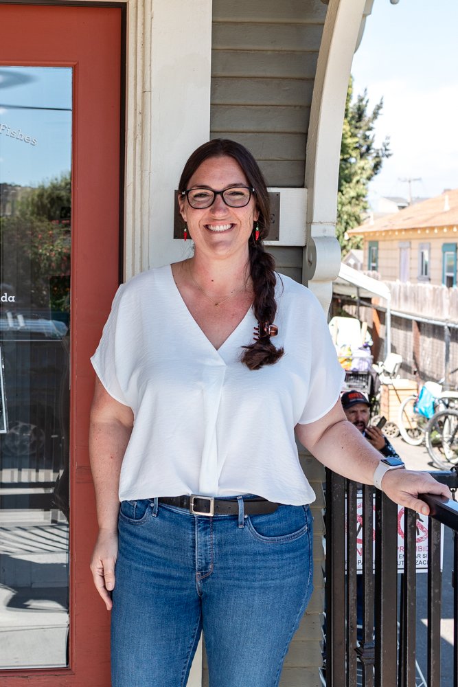 A woman with glasses and a long braid, smiling, standing outside in front of a building with a red door and gray siding.