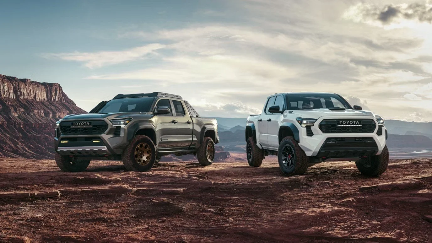 Two Toyota pickup trucks, one gray and one white, are parked on rocky terrain with a desert and canyon landscape in the background under a partly cloudy sky.