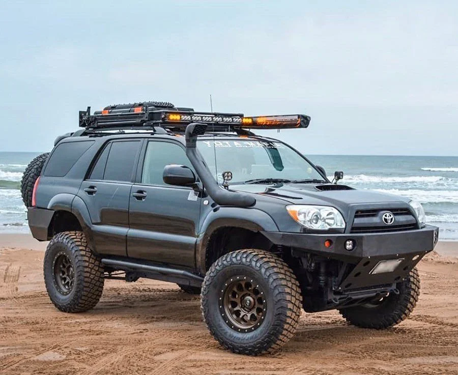 A black Toyota SUV modified for off-road driving, equipped with large tires, a roof rack, and a light bar, parked on a sandy beach with the ocean in the background.