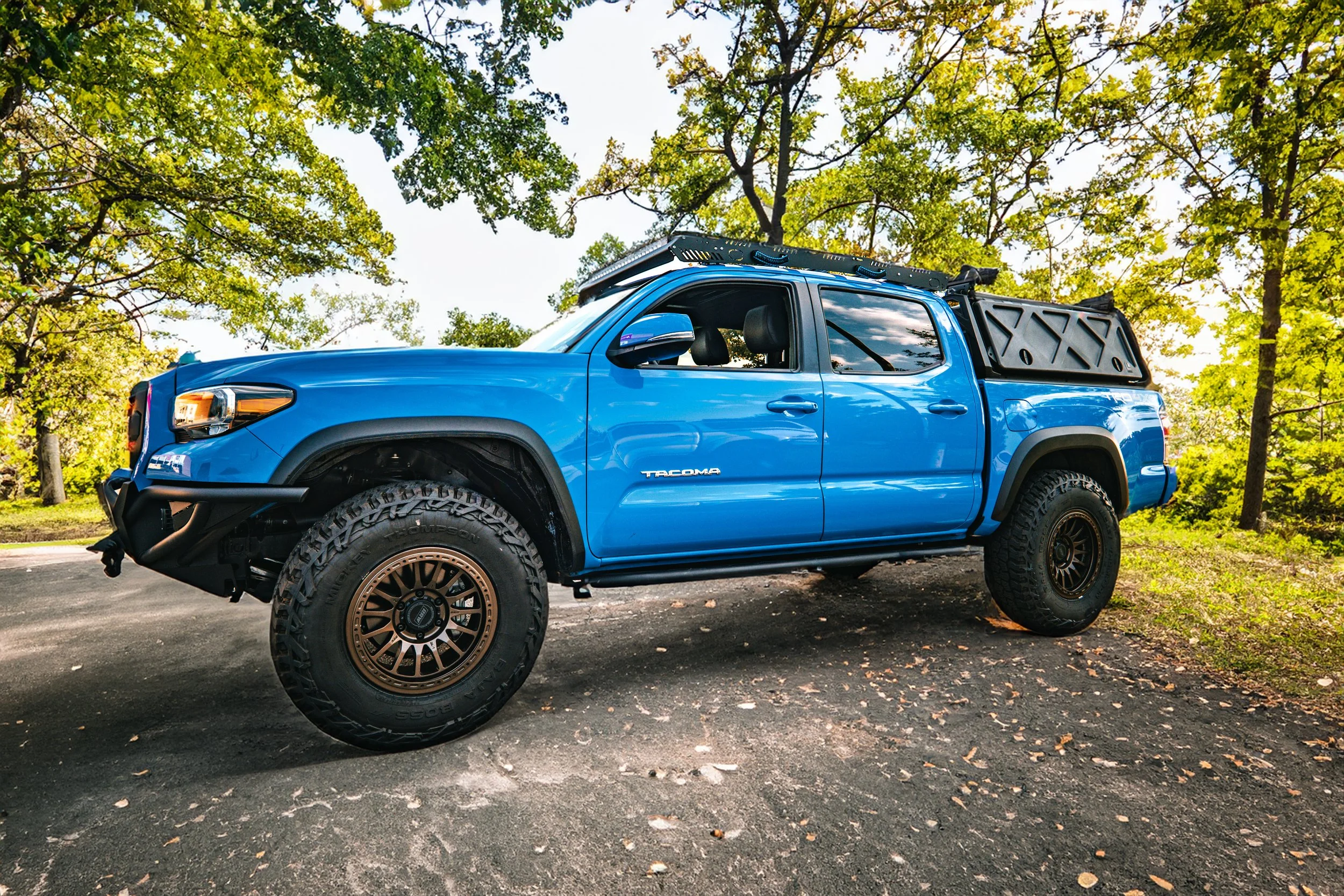 A blue Toyota Tacoma pickup truck parked on a paved surface with fallen leaves, surrounded by green trees with sunlight filtering through.
