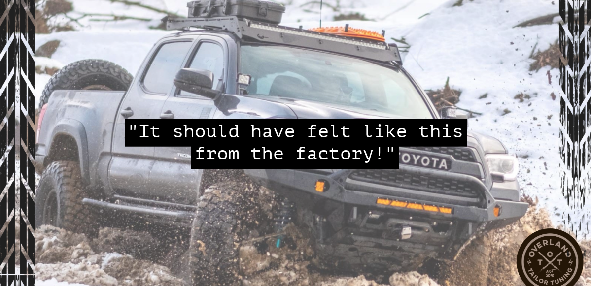 A black Toyota pickup truck driving through muddy snow, with large off-road tires and a roof rack, with a snowy landscape in the background.