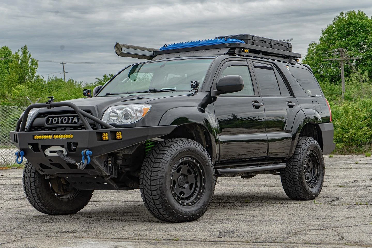 A black Toyota SUV modified for off-road use, equipped with large tires, a front bumper with a winch, auxiliary lights, a roof rack with additional gear, and a snorkel for water crossings, parked on a concrete lot with trees and a cloudy sky in the background.