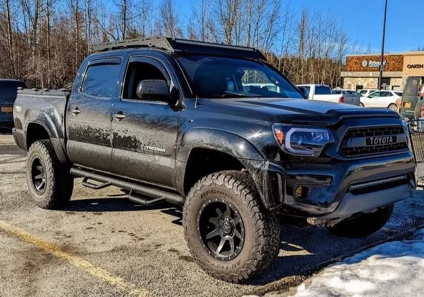 A black Toyota Tacoma pickup truck parked in a parking lot with some snow on the ground and trees in the background.