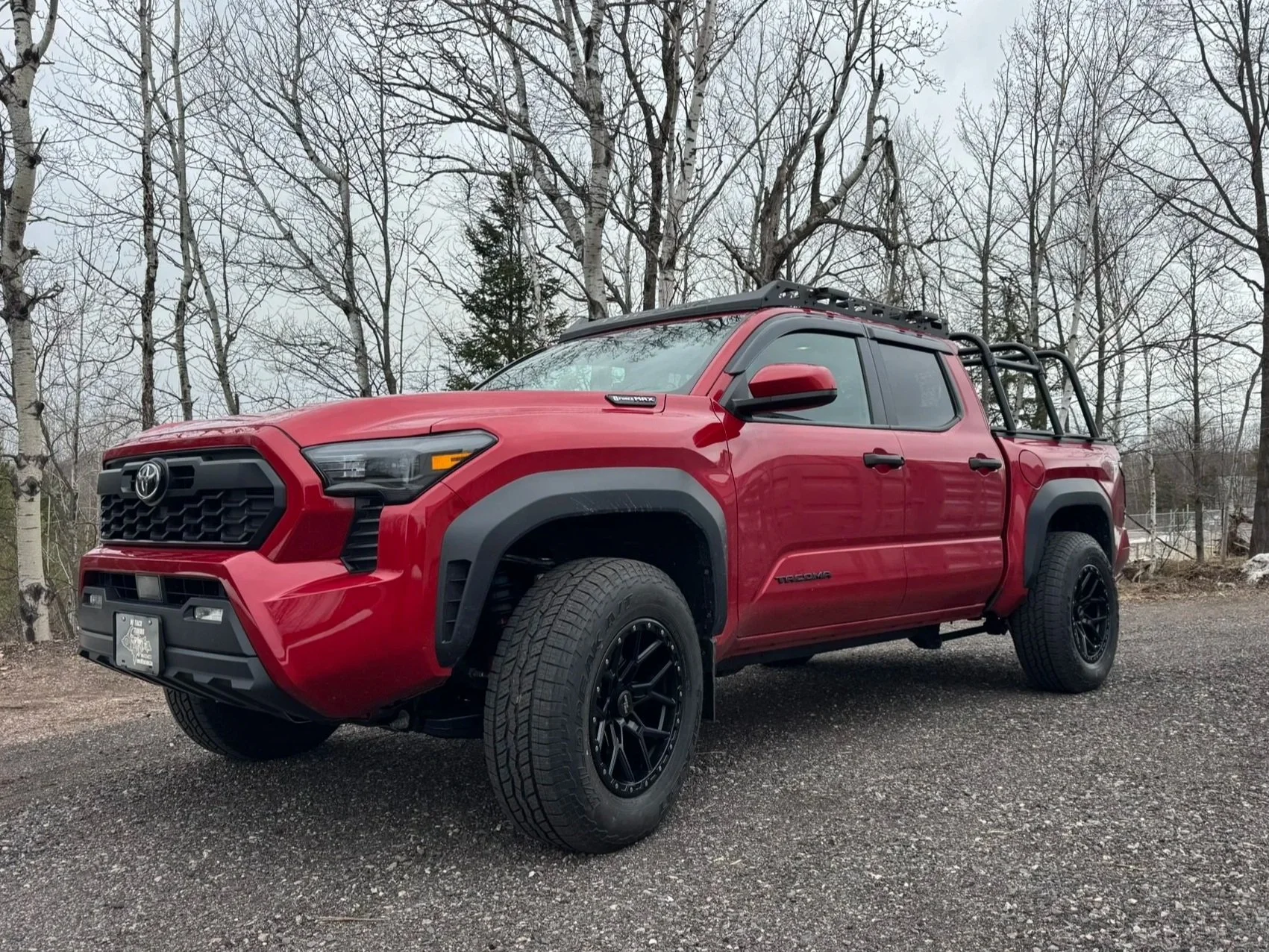 Red Toyota Tacoma pickup truck with black rims and off-road tires parked on a gravel surface, surrounded by leafless trees in a natural setting.
