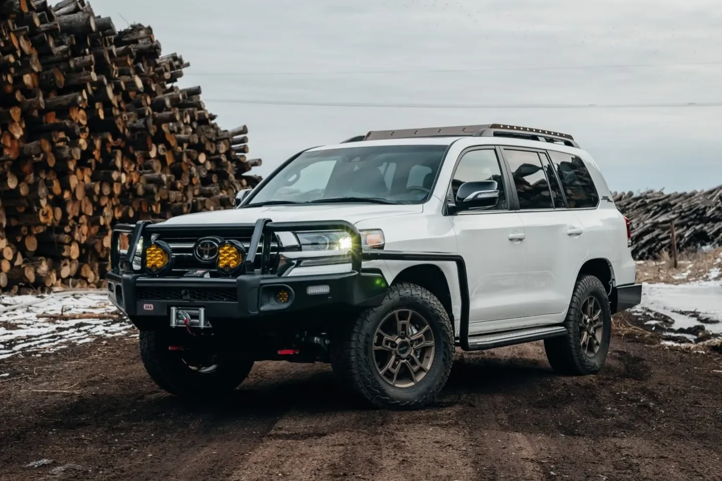 A white Toyota SUV with off-road modifications, including a heavy-duty bumper, winch, and auxiliary lights, parked on a dirt surface near a large stack of firewood.