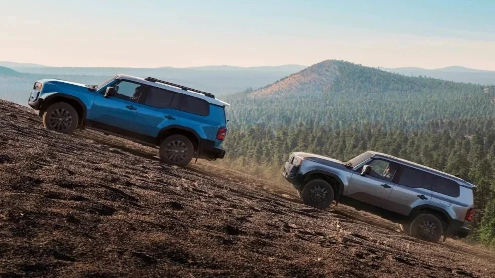 Two SUVs driving on a steep dirt incline in a mountainous area with dense forest in the background.