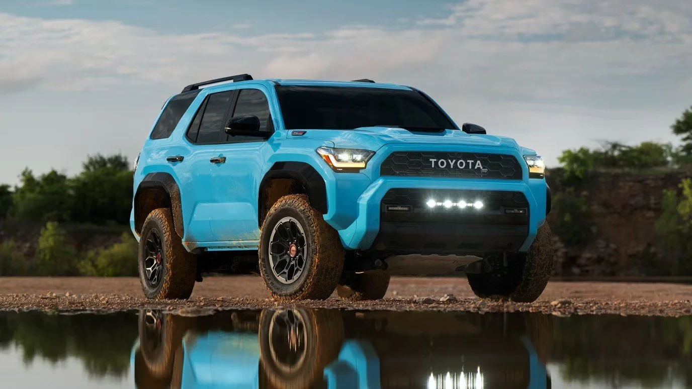 A blue Toyota SUV parked on a dirt ground near a small water puddle, with a reflection of the vehicle visible in the water, under a partly cloudy sky.