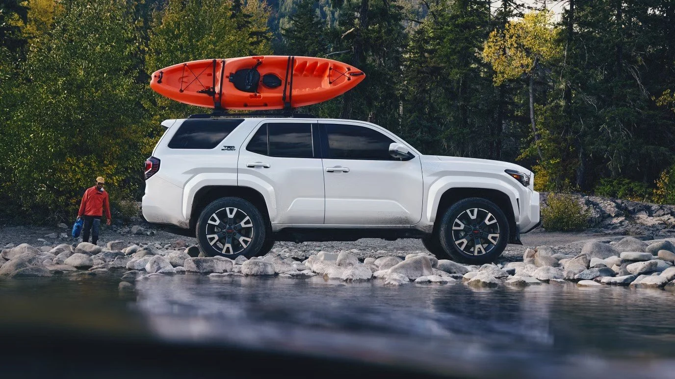 A white SUV parked on rocky riverbank with a red kayak on its roof, and a person in a red jacket and blue gloves walking nearby in a forested area.