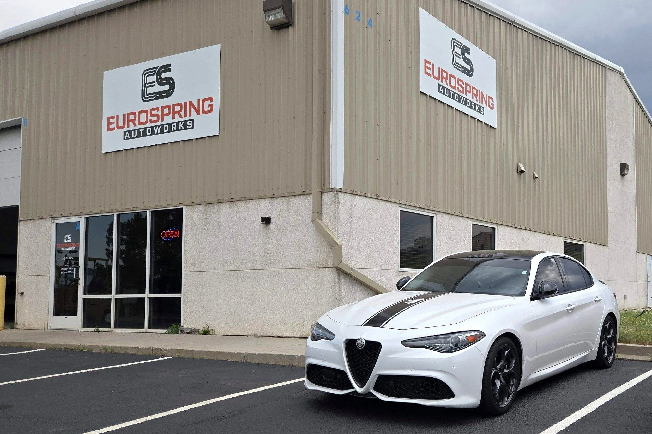 Exterior view of Eurospring Autoworks building with its logo on the wall, parked in the lot is a white Alfa Romeo