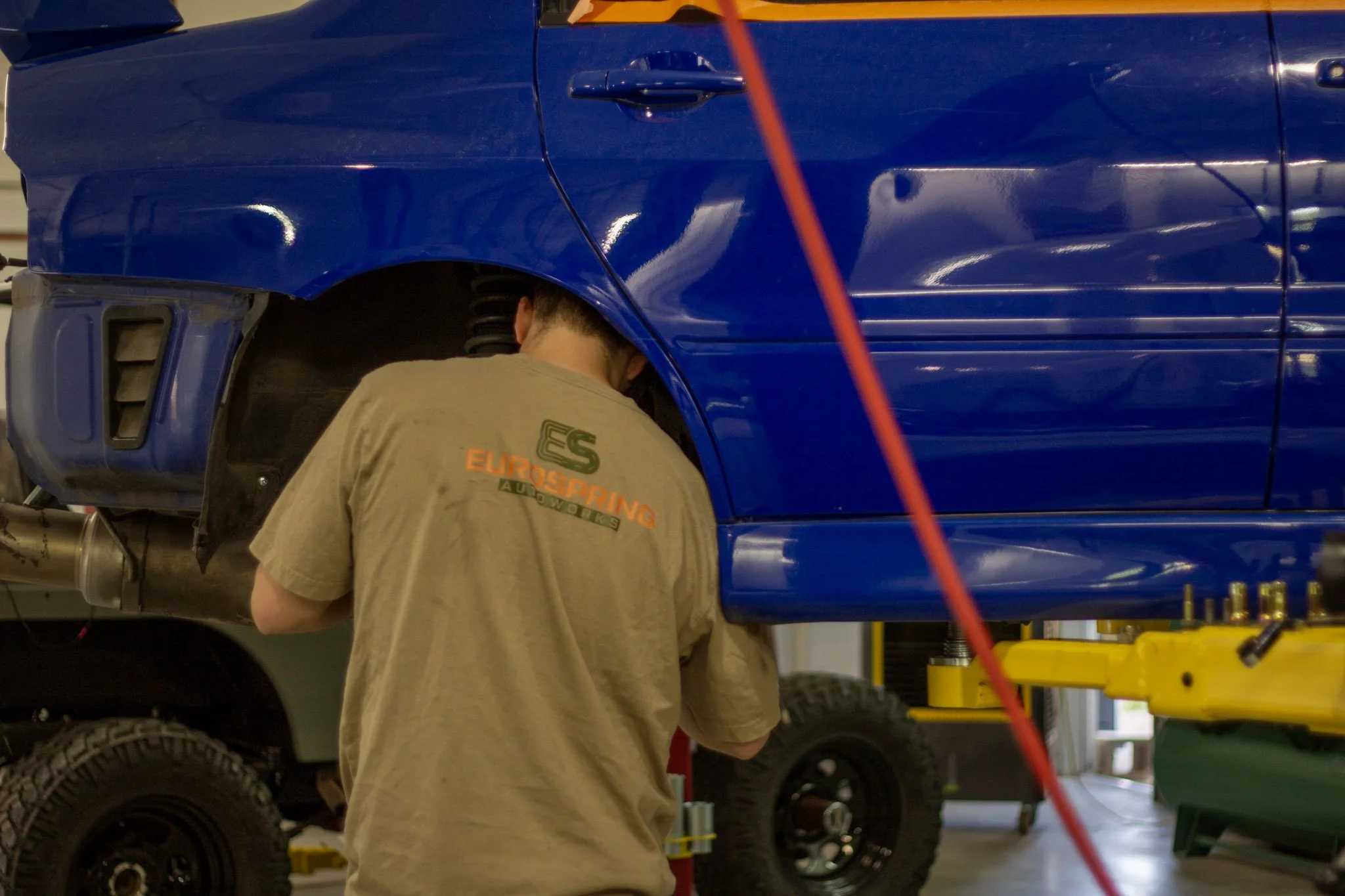 A person working on a blue vehicle in a garage or repair shop, wearing a tan shirt with orange and green text. The individual appears to be inspecting or repairing the wheel area, which is raised and supported on a stand. The garage has equipment and supplies around.