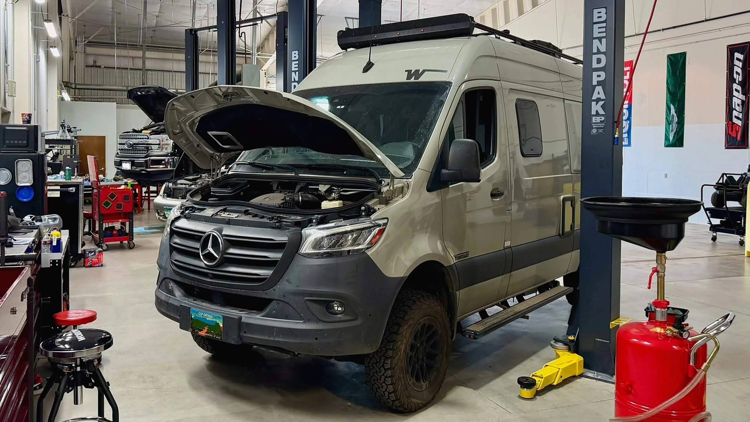 A white Mercedes-Benz Sprinter van being serviced in an indoor automotive workshop with its hood open. The workshop contains various tools, a hydraulic lift, and a black pickup truck in the background.