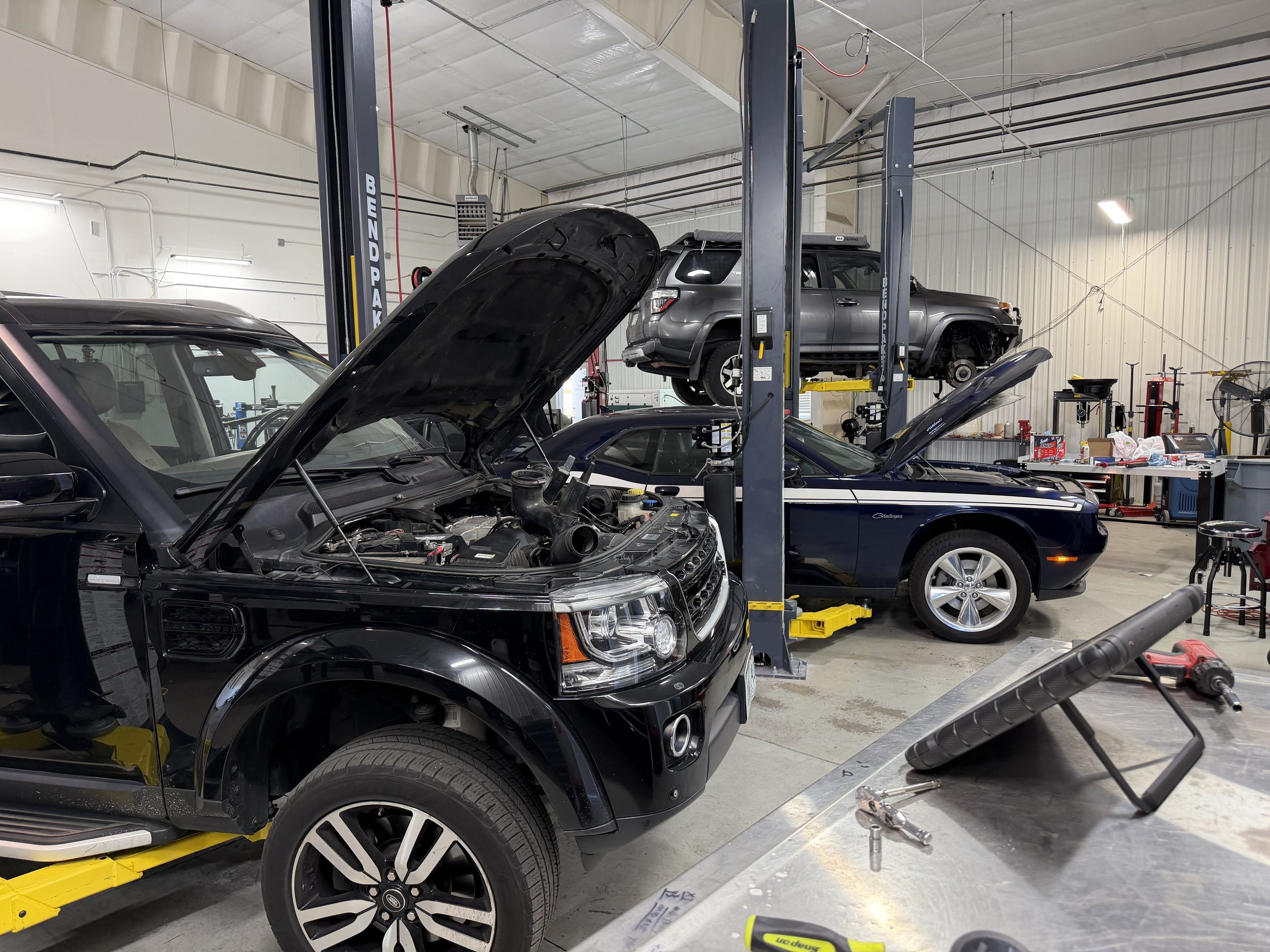 An auto repair shop with a black car having its hood open in the foreground, and a gray SUV elevated on a lift in the background. Various tools and equipment are scattered around, including a workbench to the right.