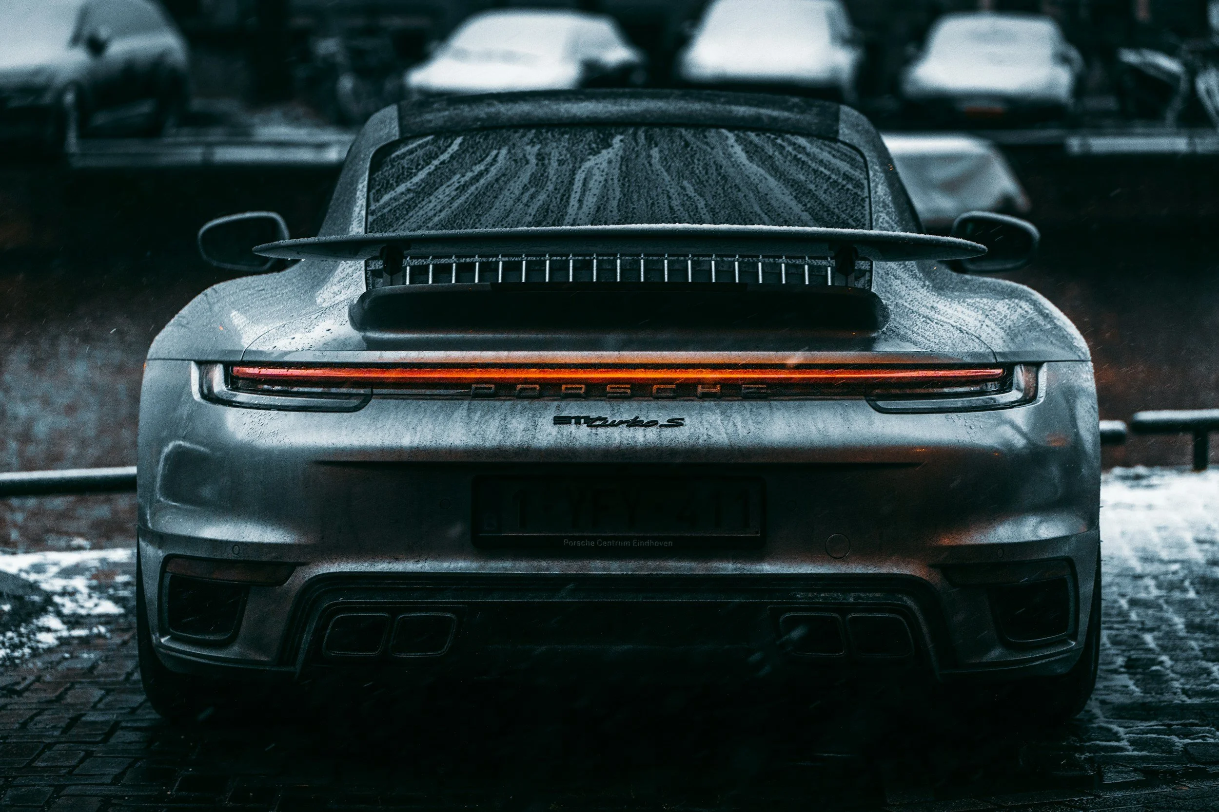 Rear view of a silver Porsche Turbo S parked on a wet brick surface, with other cars blurred in the background.
