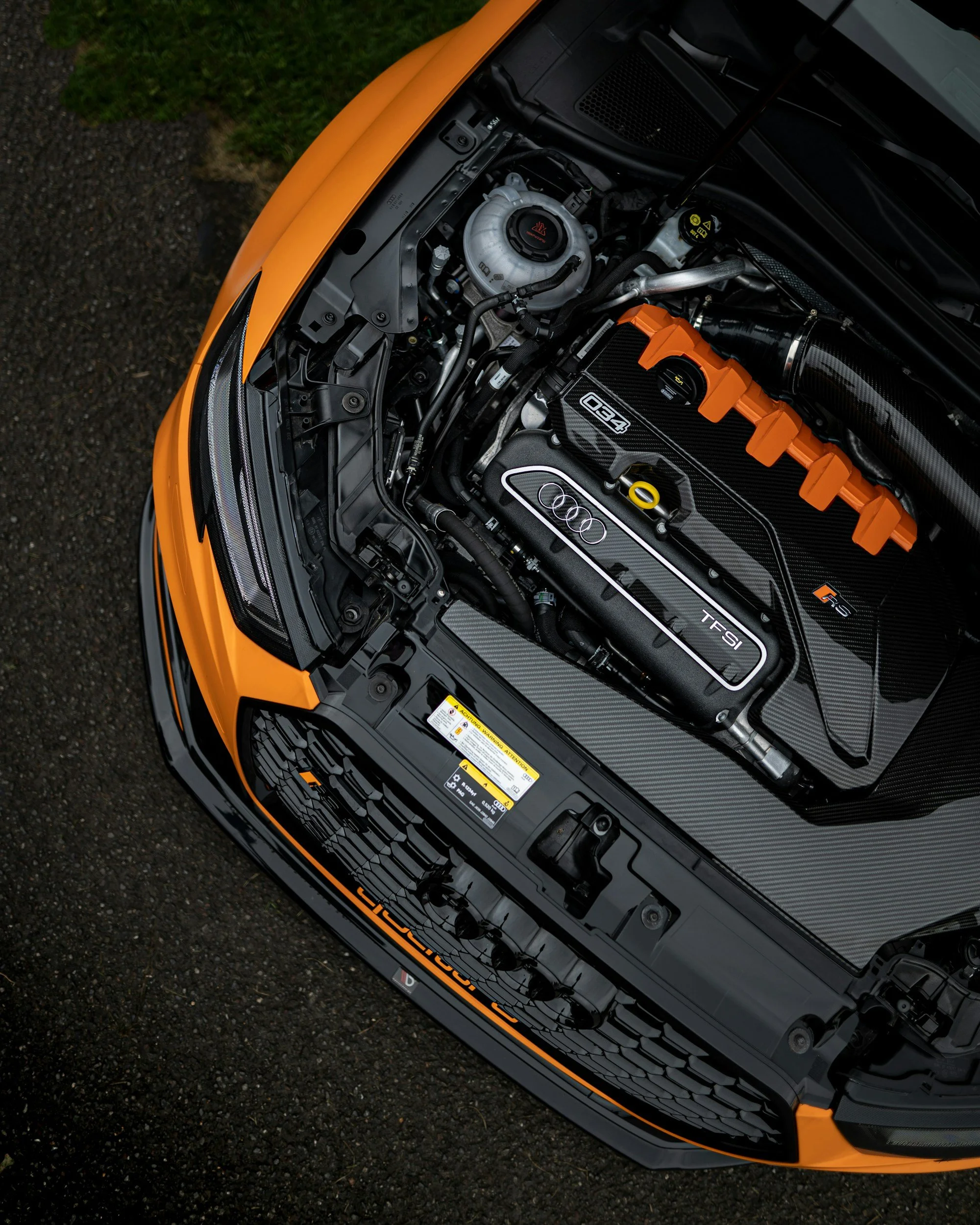Close-up of the engine bay of a yellow Audi sports car, showing the engine with black and orange accents, various hoses, components, and labels, with part of the front grille visible.