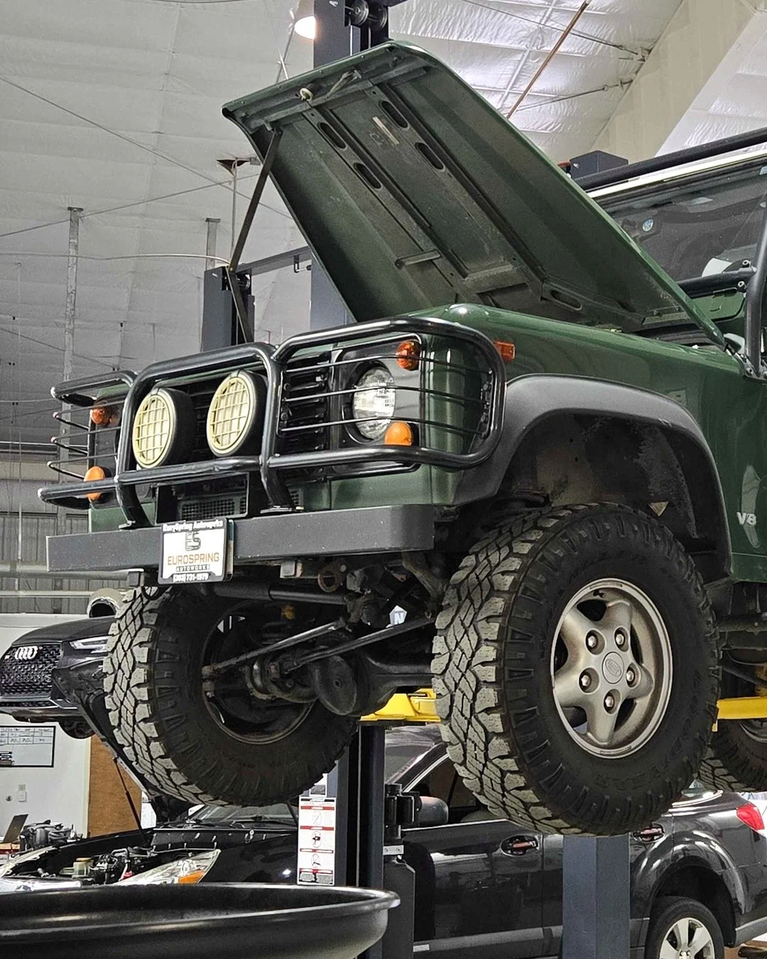 Green original Defender with its hood open, mounted on a lift in a garage. The vehicle has large tires, front grille guard, and multiple lights.