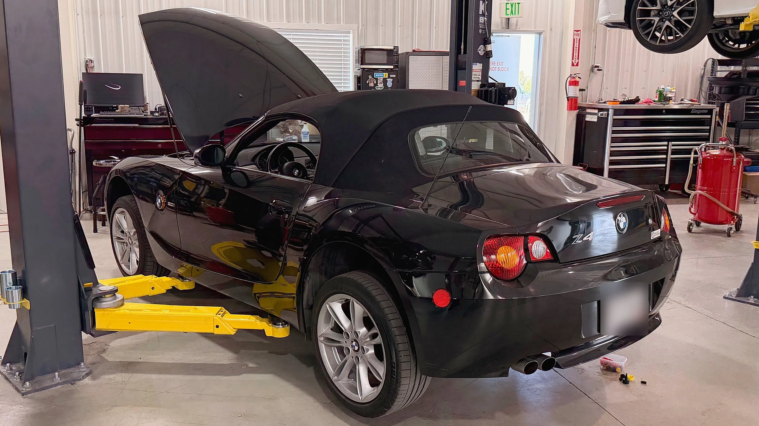 A black BMW Z4 convertible with its hood open is on a hydraulic lift in EuroSpring Autoworks. The garage contains a red tool chest, a workbench with various tools and supplies, and a fire extinguisher.