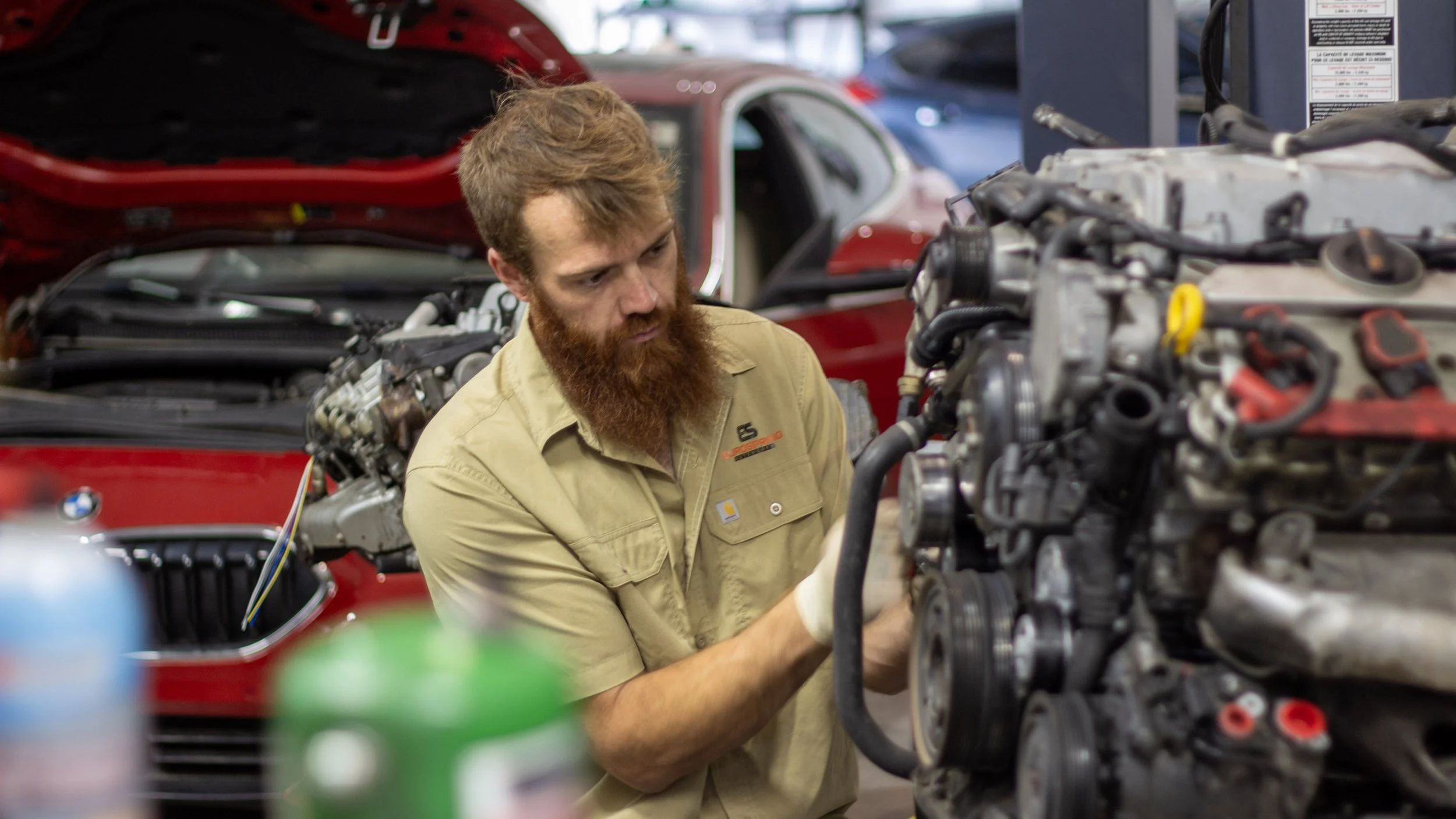 A man with a beard working on a car engine in an auto repair shop.