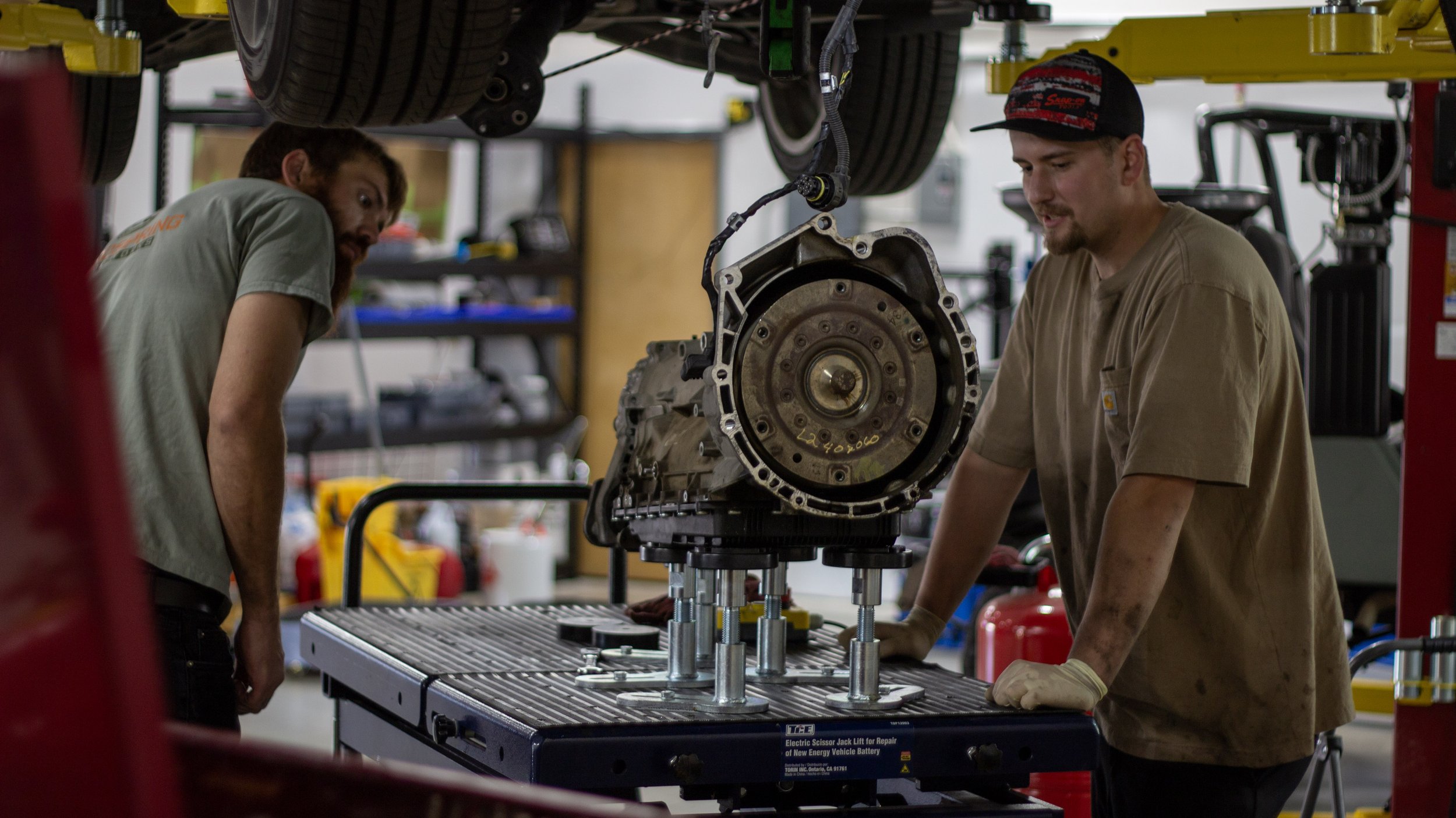 Two men work together on an automotive transmission mounted on a repair table, using a hydraulic lift to analyze or repair it in an auto repair shop.