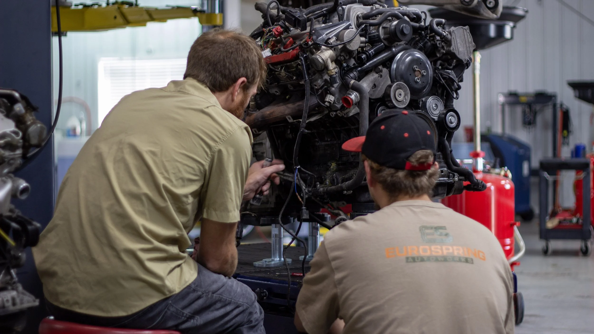 Two mechanics working on a car engine in a workshop, one kneeling and the other sitting, with various tools and equipment around.