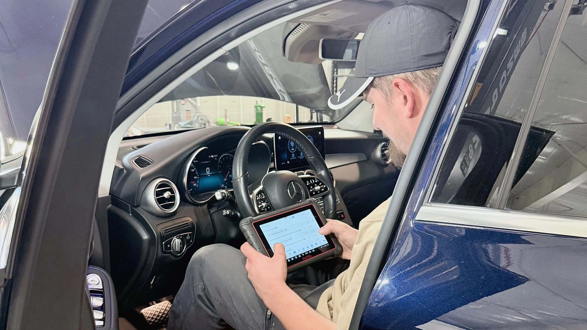 A man seated in the driver's seat of a blue Mercedes-Benz vehicle, using a diagnostic device and a tablet.