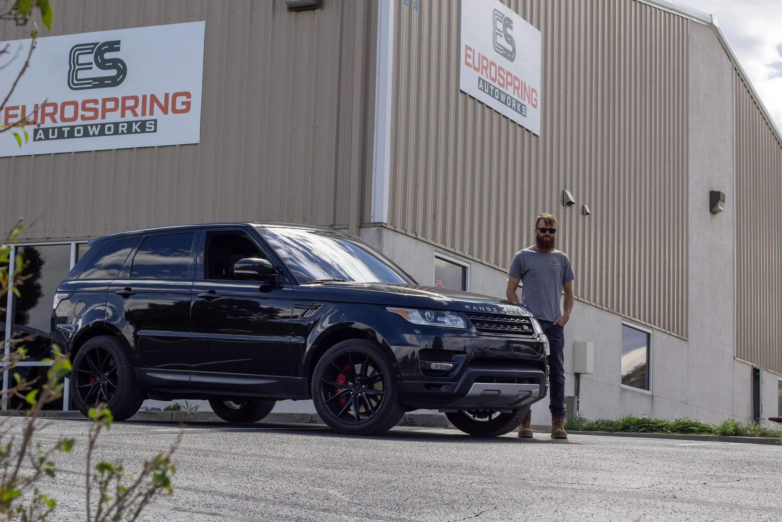 A man with a beard and sunglasses standing outdoors beside a black Range Rover parked in front of a beige industrial building with "EUROSPRING AUTOWORKS" signs.