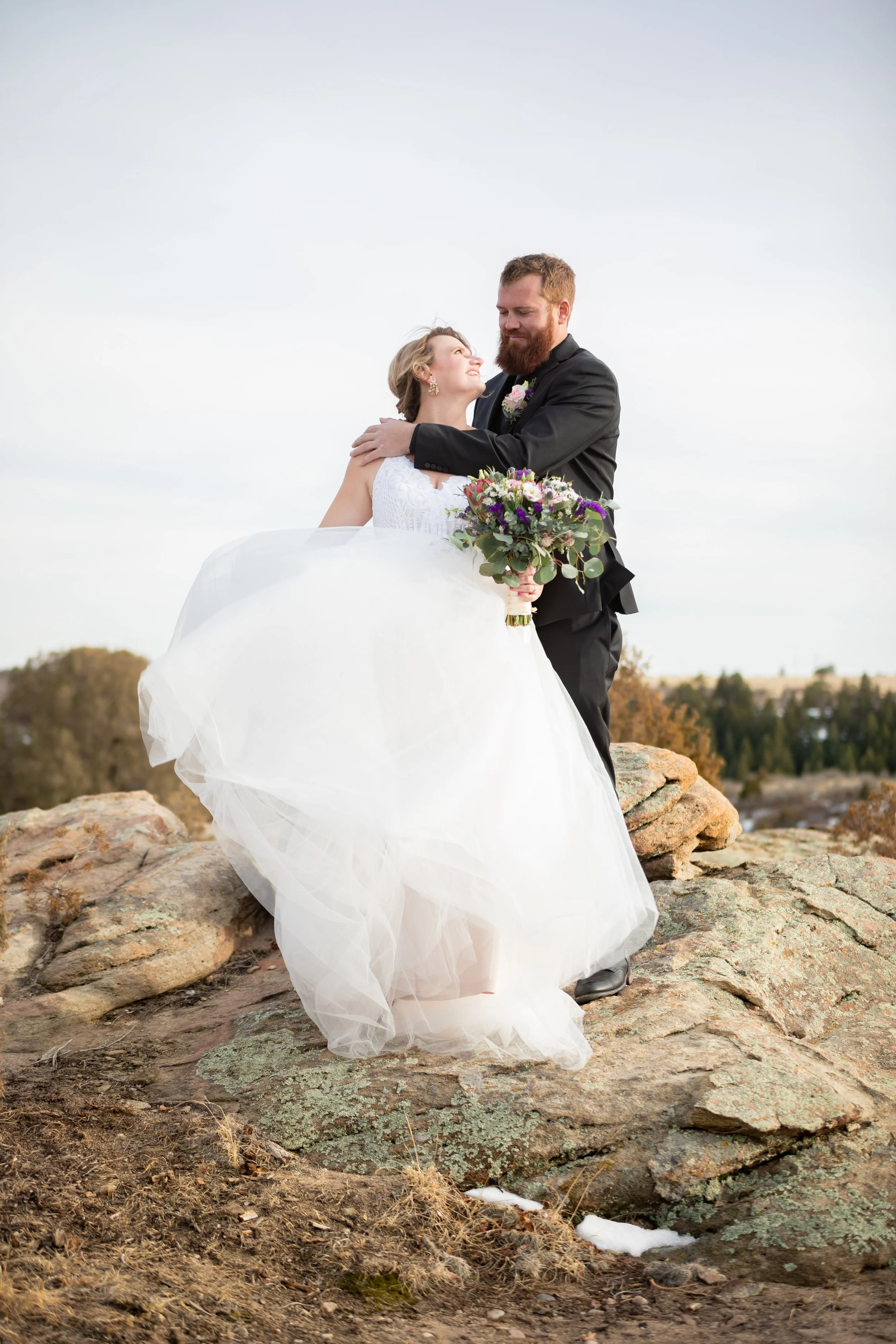 A bride and groom embracing on rocky ground outdoors, with the bride holding a bouquet of flowers, and a cloudy sky in the background.