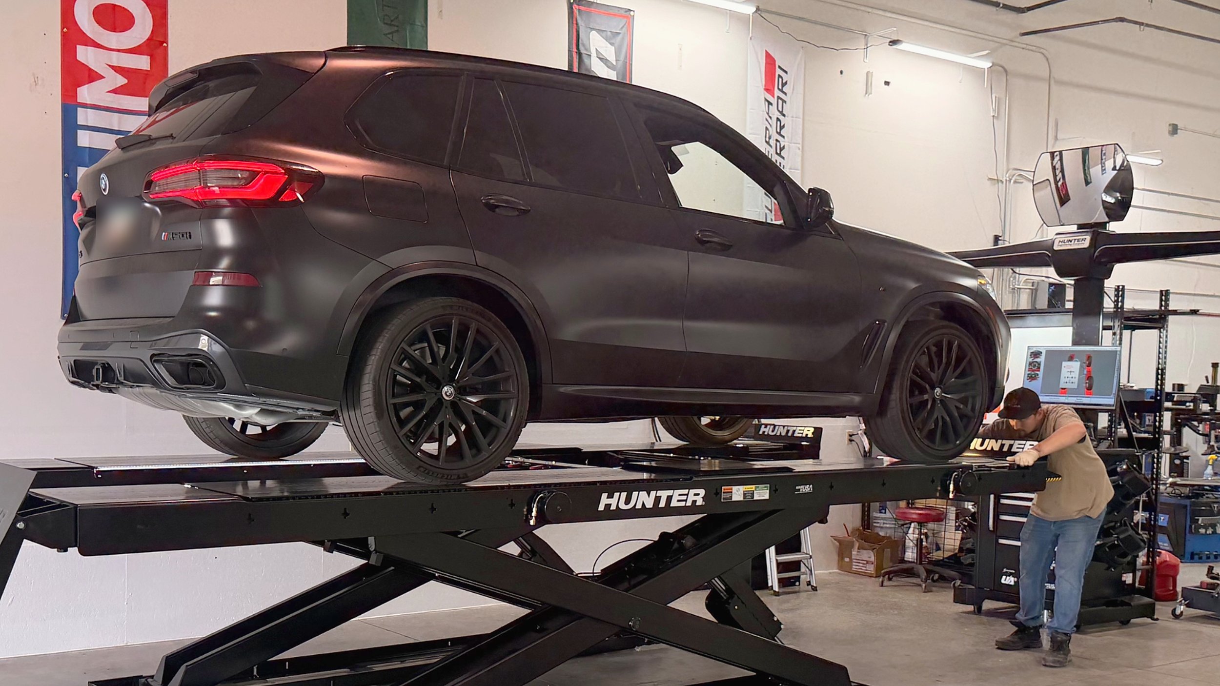 A black BMW SUV on a Hunter alignment in a workshop with banners and equipment, and a person working underneath.