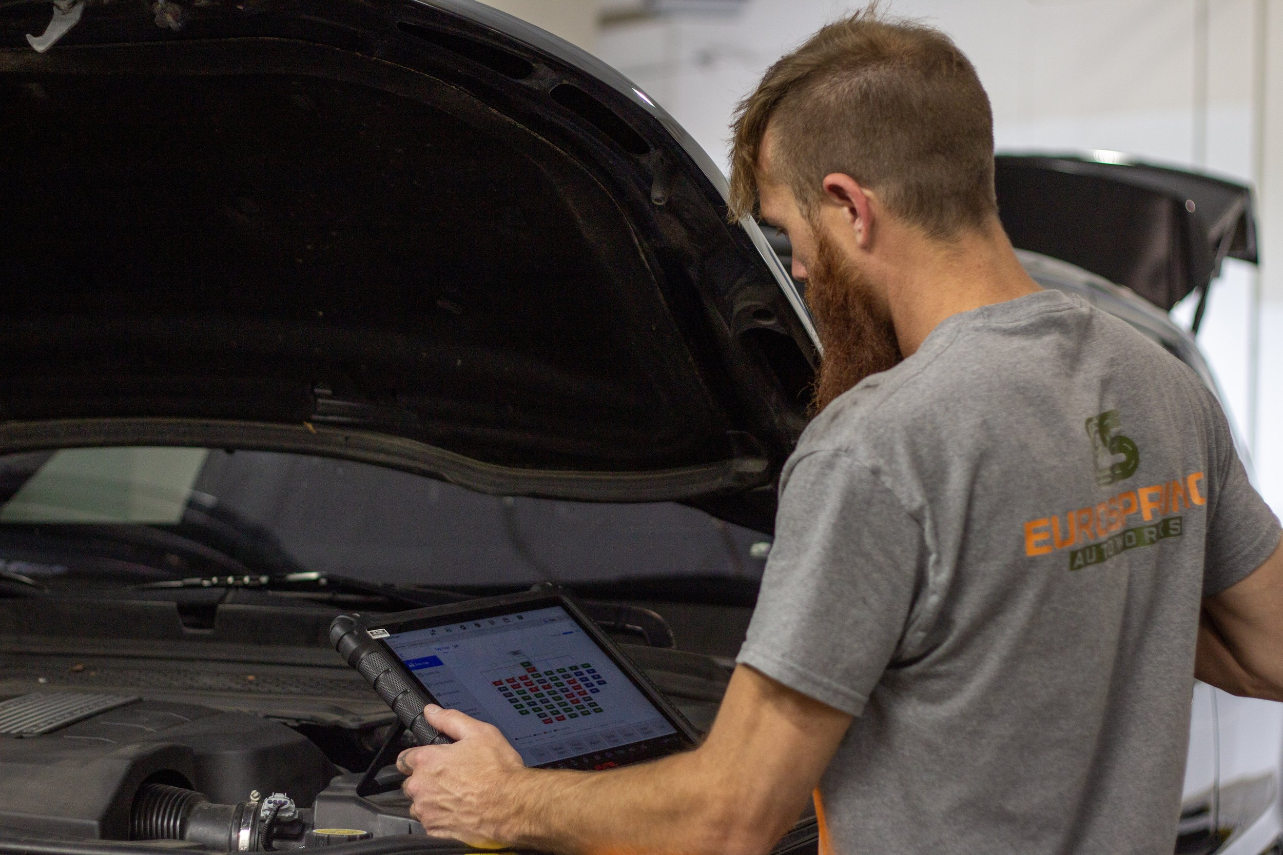 A mechanic with a beard in a gray t-shirt inspecting a car engine with a diagnostic tablet in hand.
