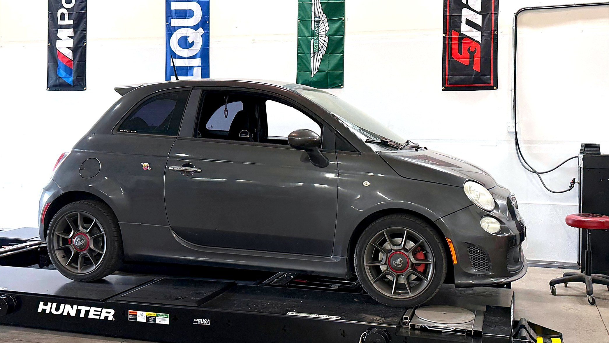 A gray Fiat 500 Abarth on the alignment rack at EuroSpring Autoworks in Castle Rock with flags hanging on the wall, including M Power, Huron, and ZRZ.