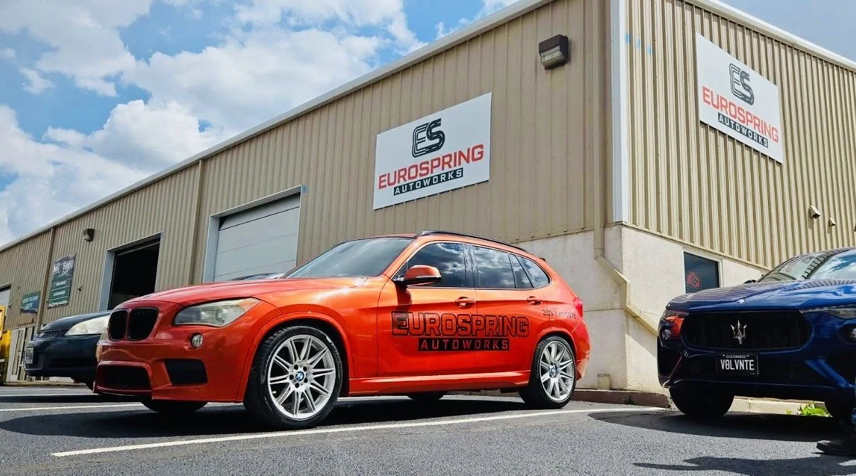 Red BMW station wagon with 'EUROSPRING AUTOWORKS' logo on side, parked in front of an auto repair shop with other cars nearby.