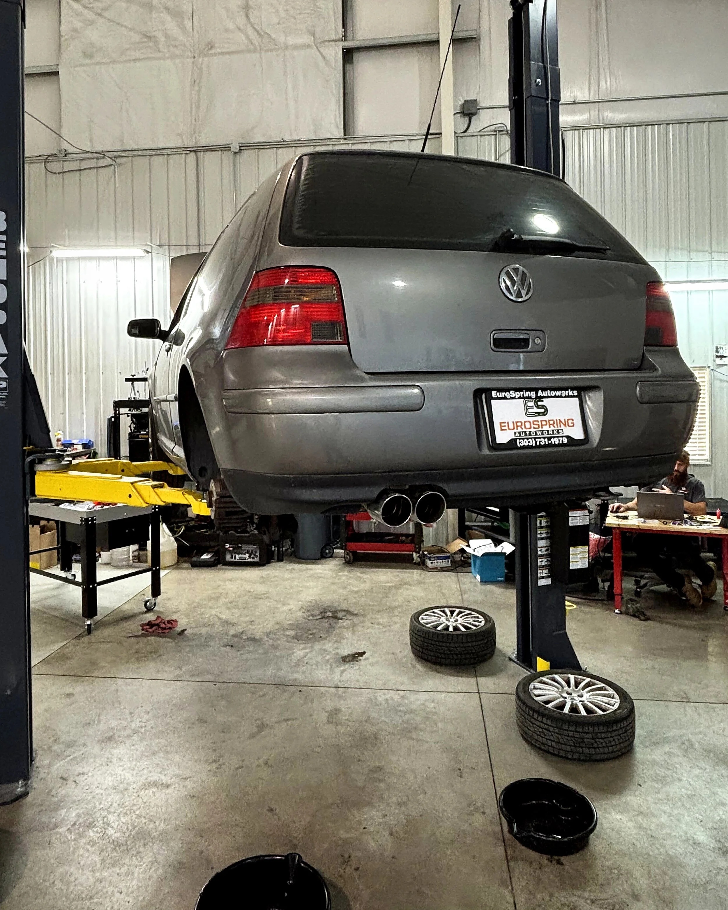 Silver Volkswagen Gold elevated on a hydraulic lift in an auto repair shop, with various car parts, tools, and equipment around. Two tires are on the floor, and a mechanic is working at a desk.